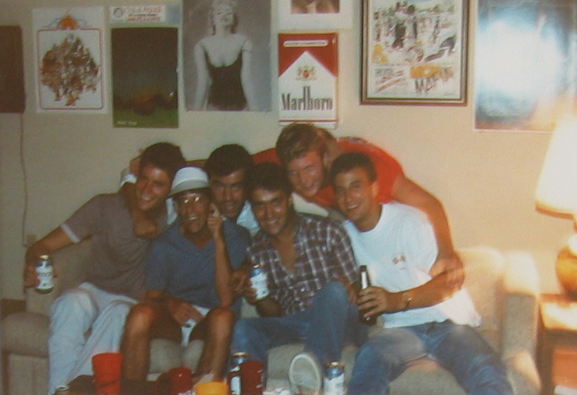 A group of six friends posing happily on a couch in a living room, holding drinks and enjoying each other's company, with colorful posters on the wall.